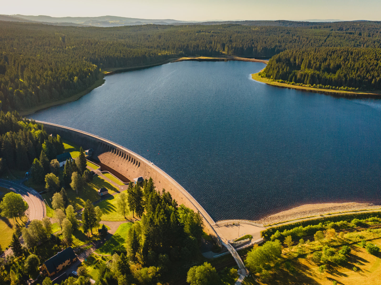 Sommerliches Luftbild mit Staumauer mittig im Bild und einen Stausee umgeben von Wald in der rechten Bildhälfte